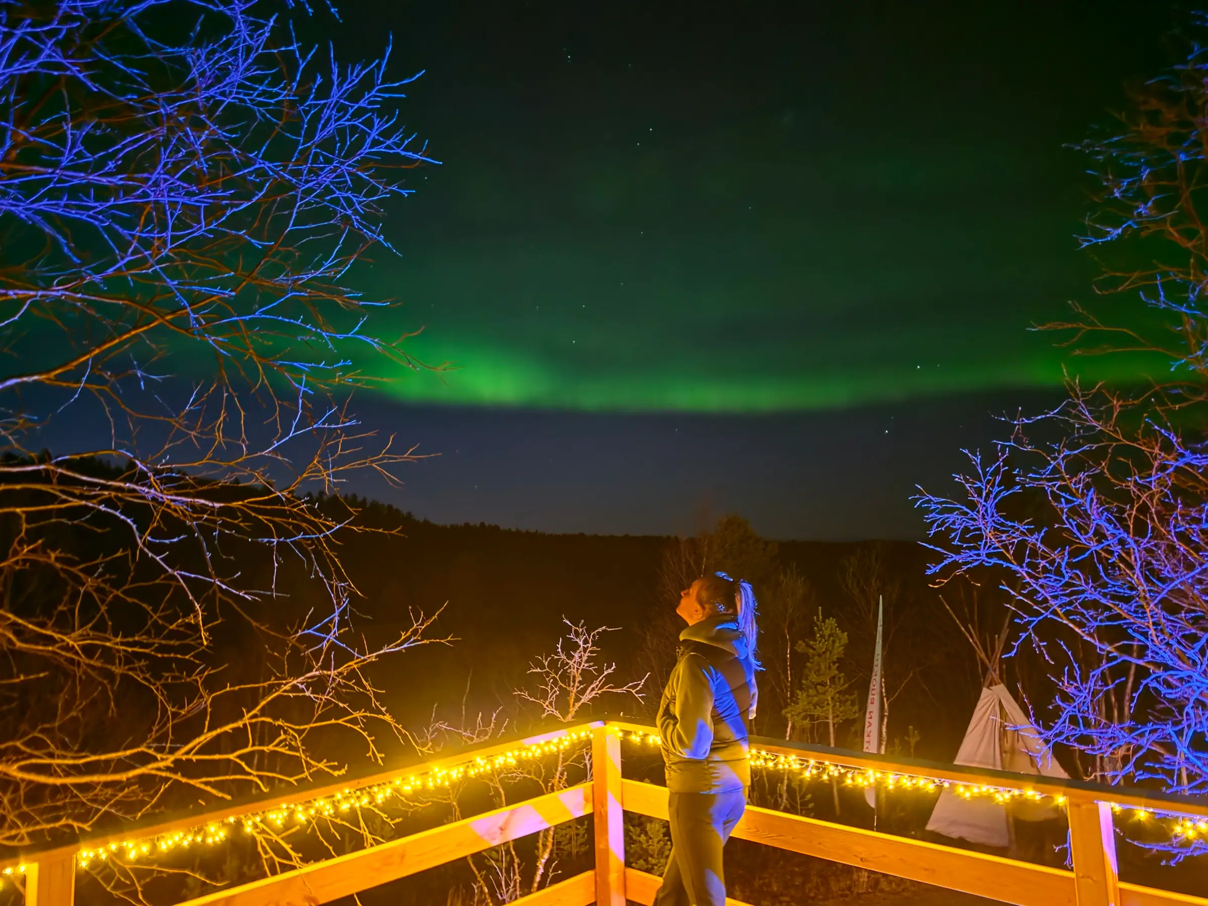 A guest on a wooden terrace in the village, looking up at the Northern Lights