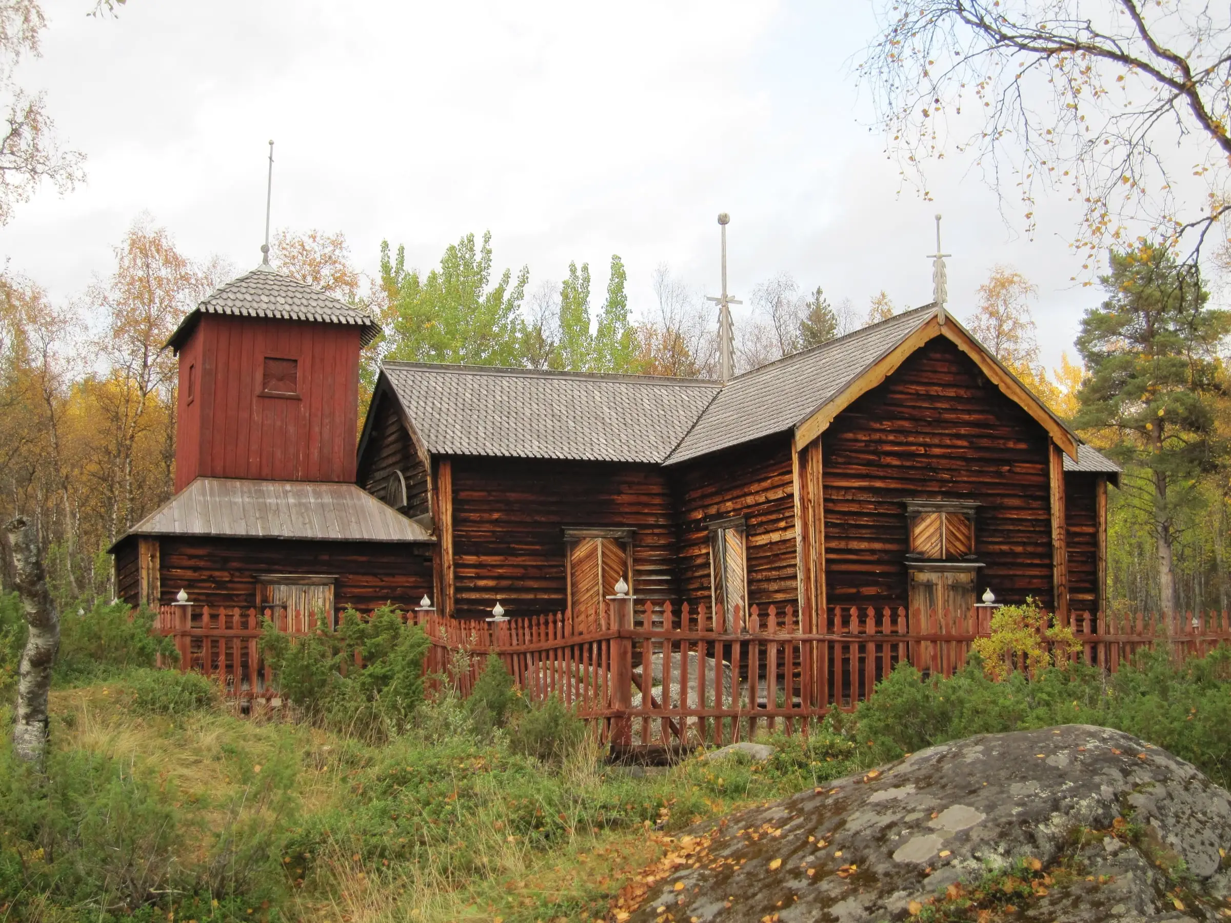 The red-brown wooden Pielpajärvi wilderness church behind a red fence, among autumn birches