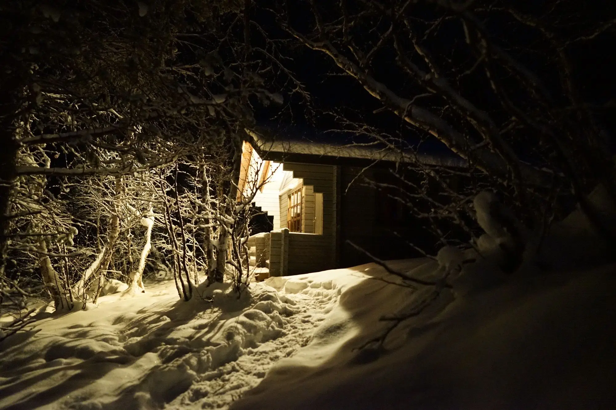 A log cabin with a warm light in its window, on a snowy path at night