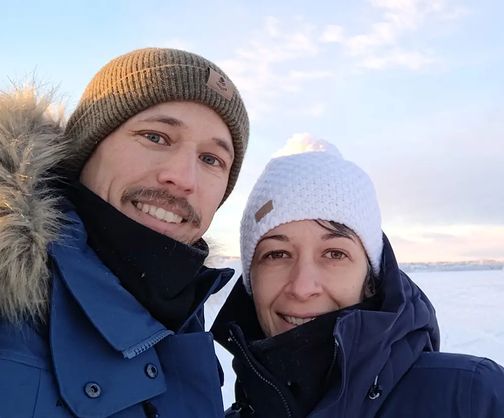 Two guests in winter jackets and woollen hats smiling in front of the frozen lake