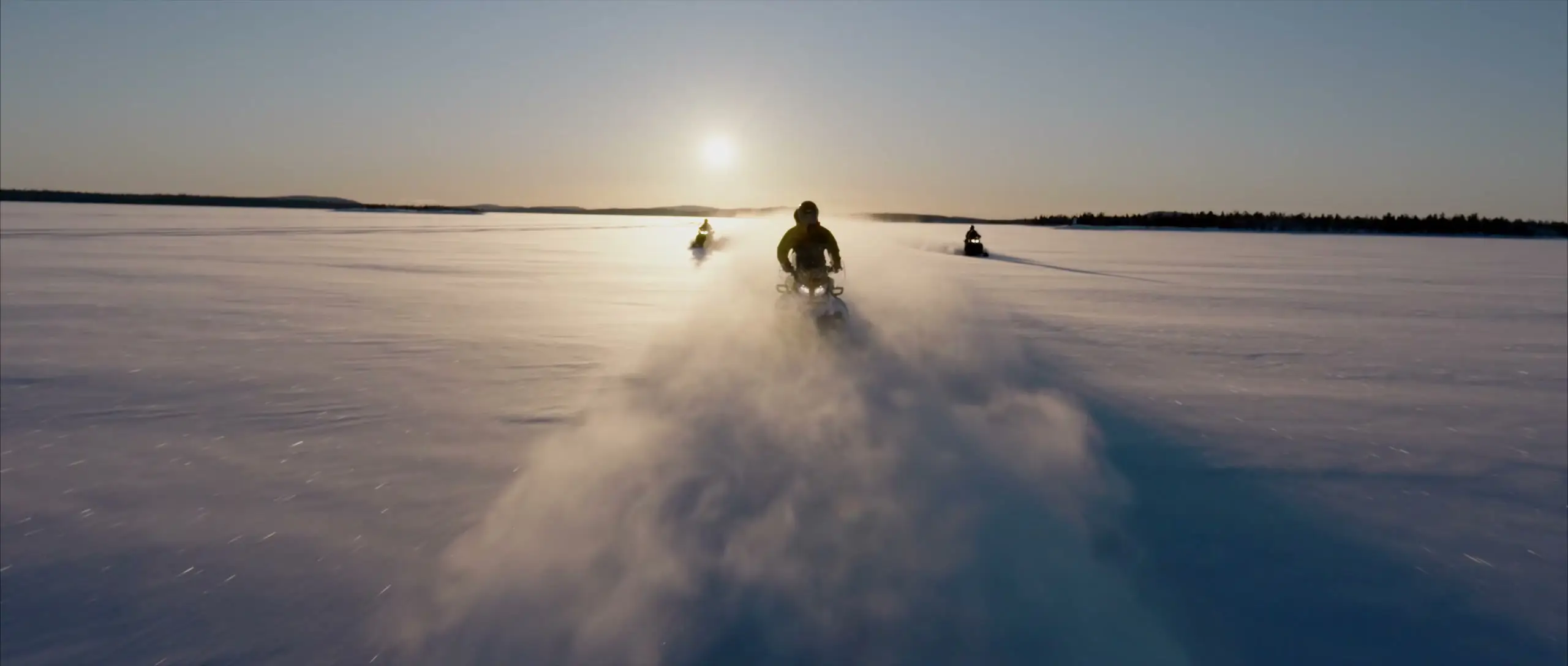 A snowmobile throws up a cloud of snow across a frozen lake towards the low sun